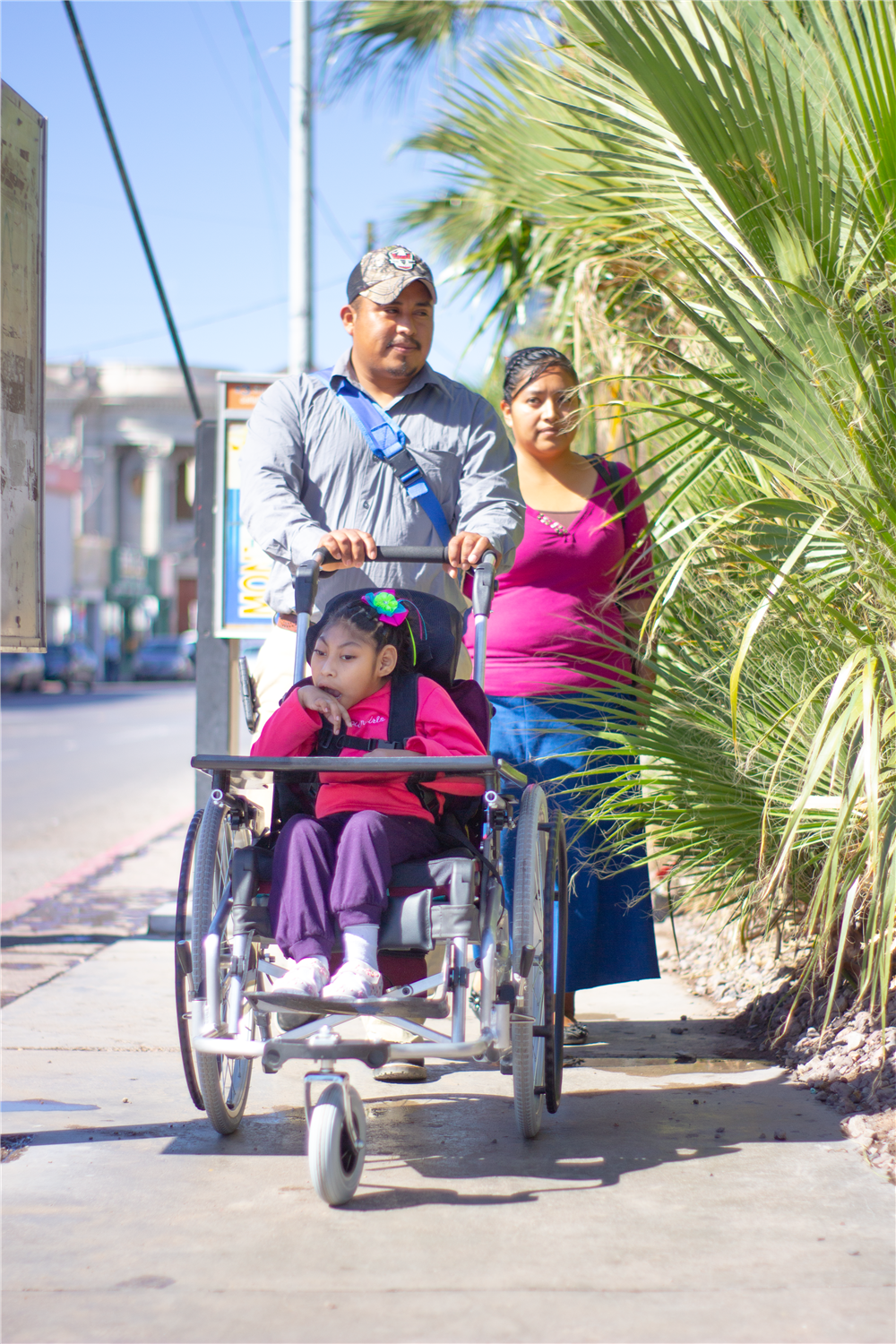 A family walks along a sunny street as a father pushes a young girl in a wheelchair, accompanied by her mother. The image gently conveys love, care, and the importance of accessible urban spaces for all families, including those with disabilities.