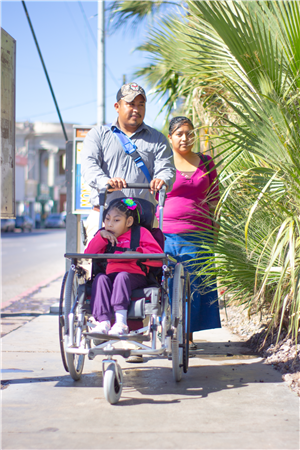 A family walks along a sunny street as a father pushes a young girl in a wheelchair, accompanied by her mother. The image gently conveys love, care, and the importance of accessible urban spaces for all families, including those with disabilities.