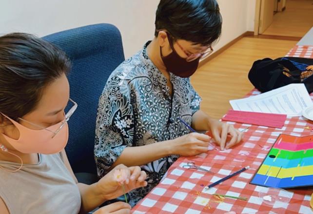 Two women wearing mask seated while doing crafts. Different colors of papers, pencil and a pair of scissors are on the table.