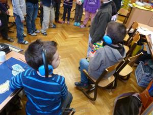 Two boys with blue headphones sit on chairs, while the rest of children are standing in a circle.