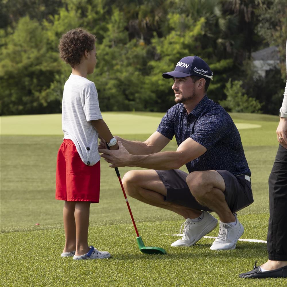 On a green golf course, an adult kneels to guide a child holding a club. The scene reflects patience, mentorship, and inclusion through sport, emphasizing how shared activities build confidence and bridge generations with care and respect.