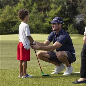 On a green golf course, an adult kneels to guide a child holding a club. The scene reflects patience, mentorship, and inclusion through sport, emphasizing how shared activities build confidence and bridge generations with care and respect.