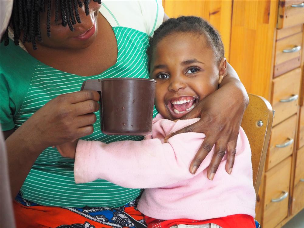 This is a heartwarming photo showing a moment of care and connection between two individuals. A woman, likely of African origin, is gently holding a mug for a young child in her arms. The child, smiling broadly and looking directly at the camera, appears to be very happy and comfortable in the woman's embrace. The setting seems to be a home environment, with wooden furniture and cabinetry in the background. The image conveys themes of nurturing, support, and the joy found in simple acts of kindness. The woman's action exemplifies assistance and the child's joyful expression reflects the positive impact of such care.