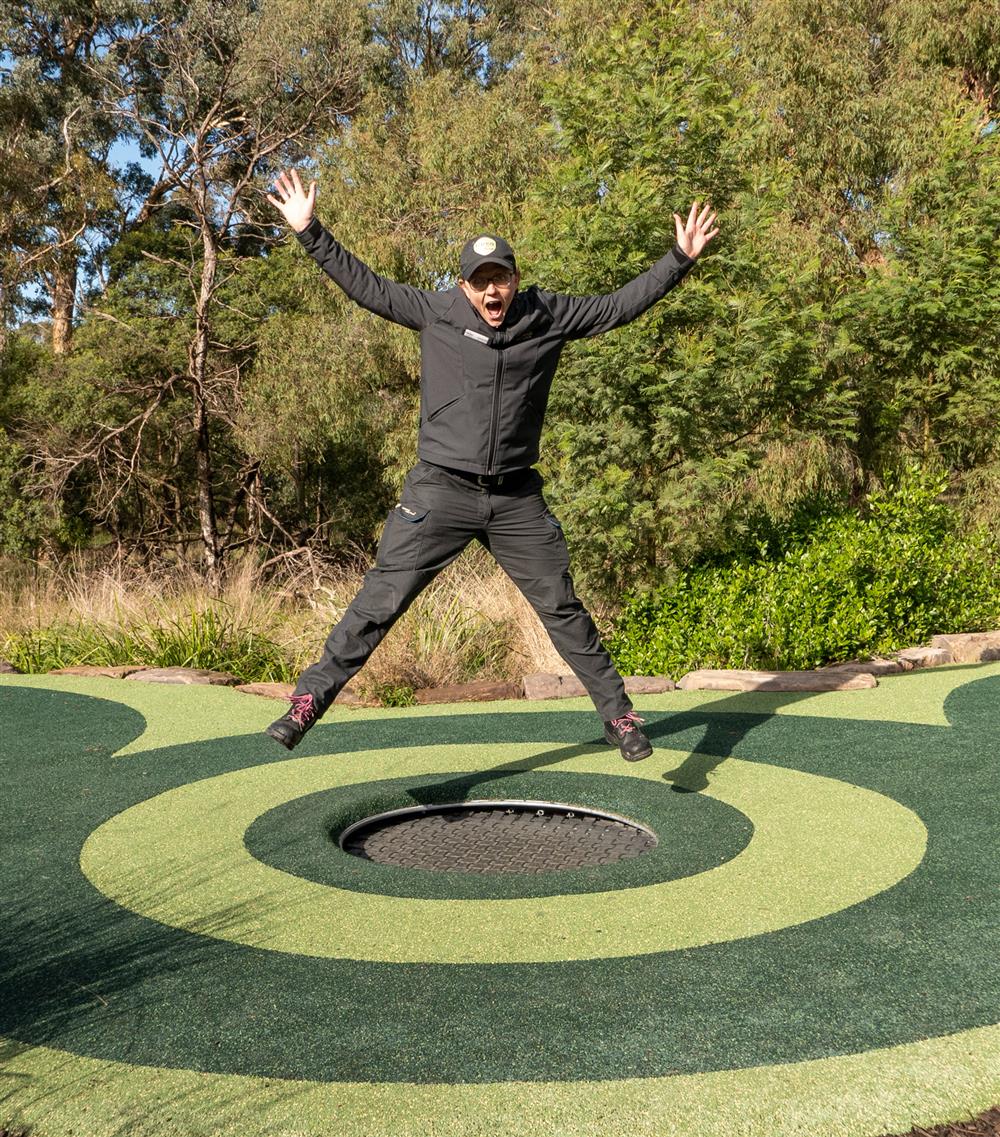 A man joyfully jumps on an outdoor trampoline surrounded by greenery. His open arms and bright expression capture the spirit of freedom and inclusion in public recreational spaces designed for everyone to enjoy safely.
