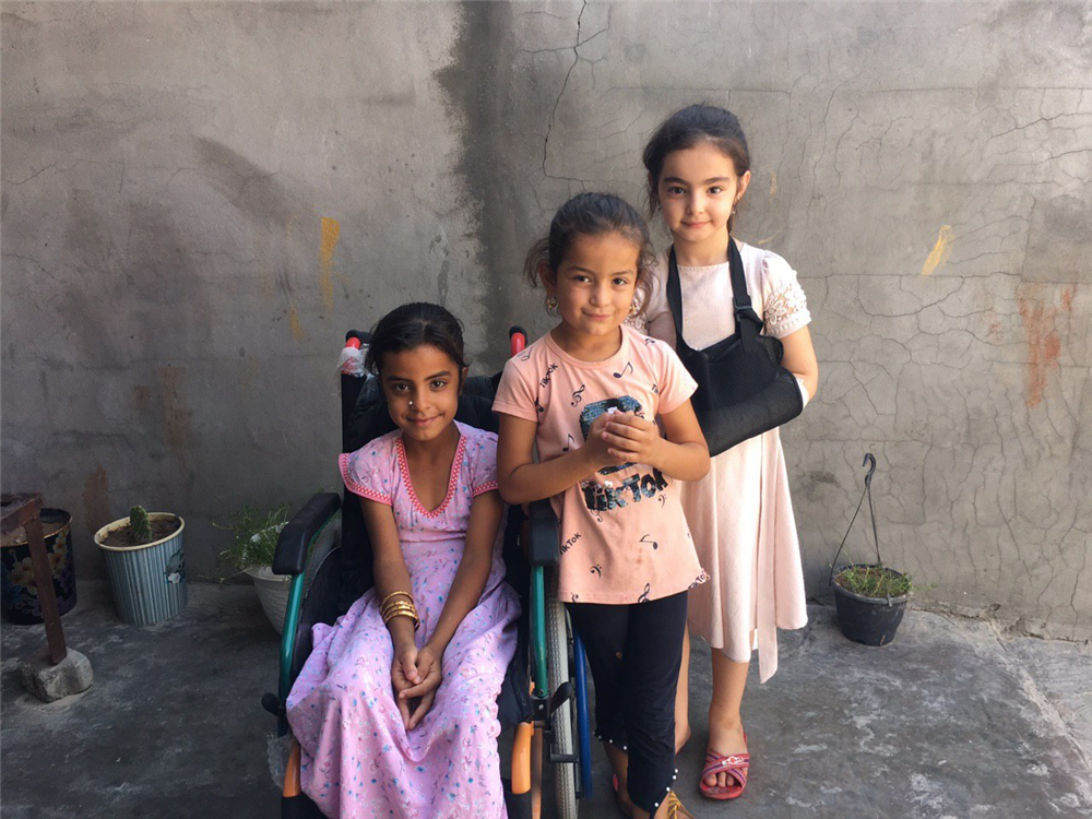 Three young girls smile into the camera while standing in front of a grey concrete wall interspersed with fine cracks. The girl on the left is sitting in a wheelchair while the girl on the far right has her arm stabilized in a sling. 