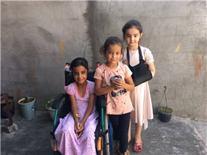 Three young girls smile into the camera while standing in front of a grey concrete wall interspersed with fine cracks. The girl on the left is sitting in a wheelchair while the girl on the far right has her arm stabilized in a sling. 
