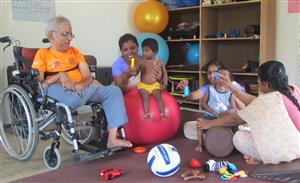 A man sits in a wheel chair, while women are supporting children on round objects and holding colorful stripes during the therapy session at the early intervention centre.