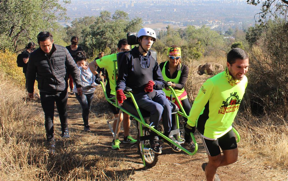 Accompanied by fellow hikers, two men and one women are pushing a single-wheel transport vehicle carrying a man with a physical disability up a sandy hiking trail.