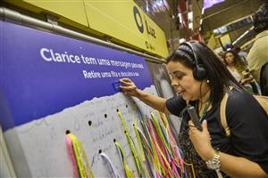 A woman interacts with a colorful public installation in a subway station, using braille and audio tools. The project, written in Portuguese, invites people to discover messages—celebrating accessibility, communication, and shared human connection.