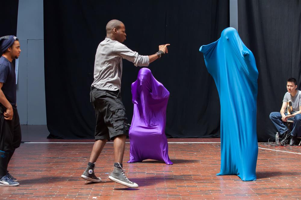 A man is acting in the theater stage with two persons fully covered with blue and purple stretchable fabric while two other men watching from a distance.