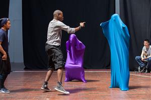 A man is acting in the theater stage with two persons fully covered with blue and purple stretchable fabric while two other men watching from a distance.