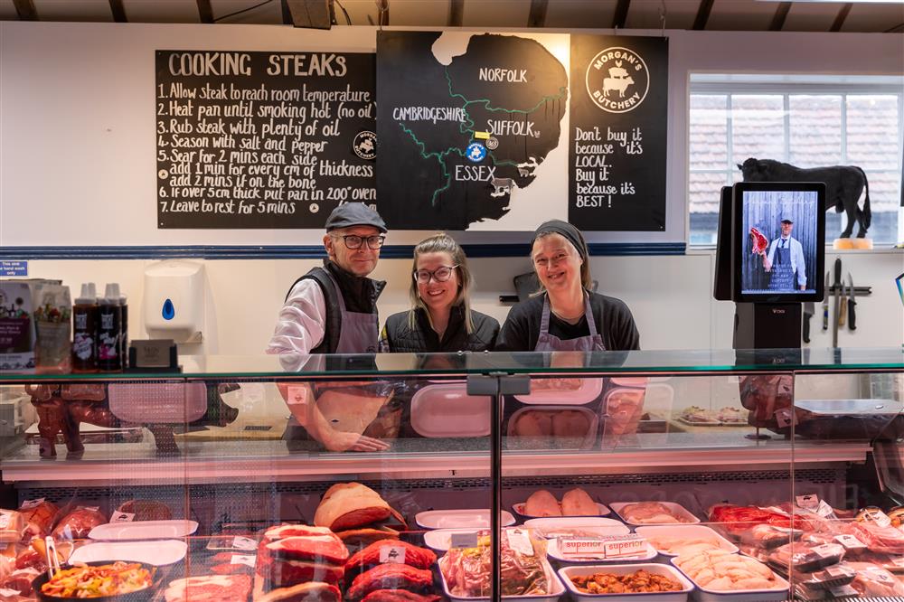 Three smiling butchers stand proudly behind a counter filled with fresh produce in a local shop. Their teamwork and welcoming atmosphere highlight the value of community-based businesses built on inclusion, cooperation, and mutual respect.
