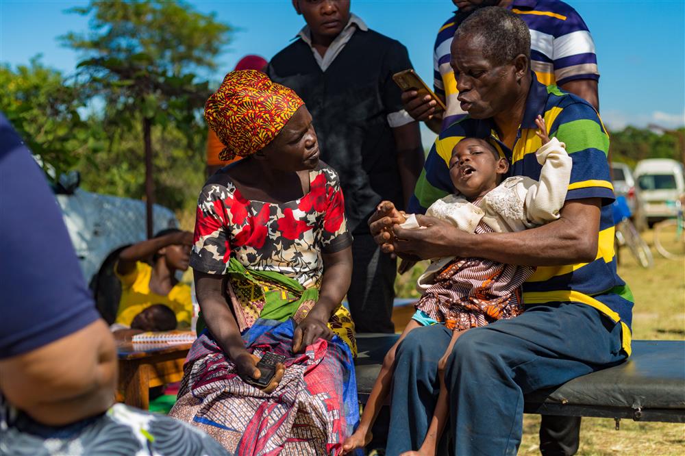 An African family sits outdoors; a father gently holds a child with a physical disability while speaking with the mother. Their interaction radiates warmth, care, and resilience—reflecting love, dignity, and the strength of family support in the face of challenges.