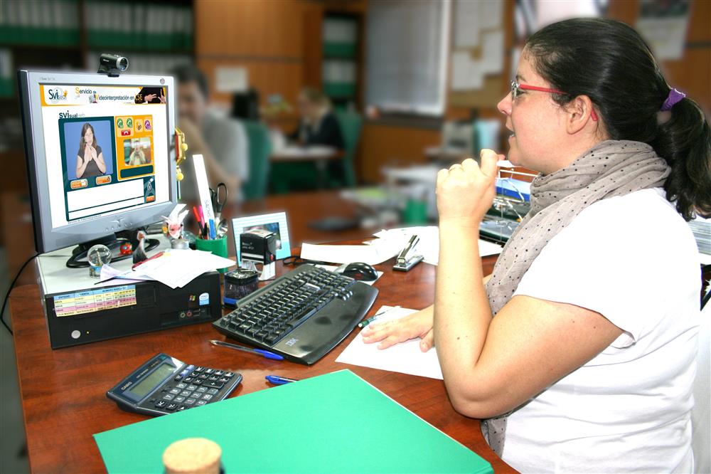 A women in a white shirt and glasses sits in a library in front of a screen talking to another person by using an online video interpretation service called SIVisual. 