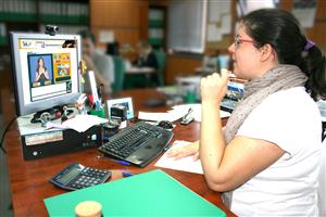 A women in a white shirt and glasses sits in a library in front of a screen talking to another person by using an online video interpretation service called SIVisual. 