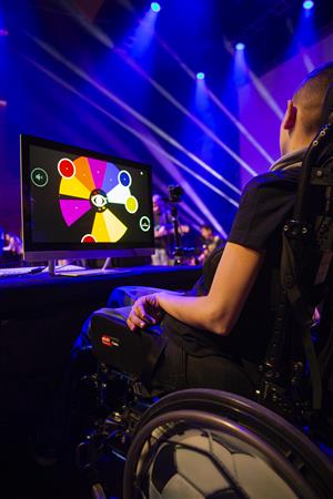 A person using a wheelchair participates in an interactive digital event, illuminated by colorful stage lighting. The image celebrates how technology and creativity can empower people of all abilities to engage fully in social and cultural experiences.