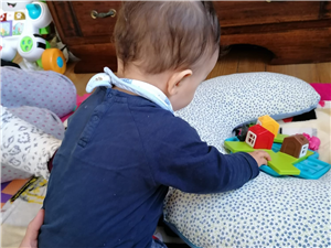The photo shows a young child from the back, engaged in play with colorful toy blocks that resemble little houses. The child is sitting on a patterned blanket that covers the floor, suggesting a safe and comfortable play environment. The presence of toys indicates a nurturing setting that encourages development and learning. The image reflects themes of growth, potential, and the universal aspect of childhood, where play is a common language across cultures, promoting equality and understanding among the youngest members of our global community.