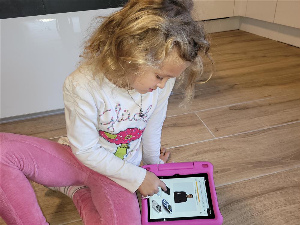 This image shows a young child engrossed in using a tablet. The child is sitting on the floor with a focus that suggests engagement with the content on the screen. The tablet is encased in a protective pink cover, indicating that it may be specially designed for use by children. The child appears to be casually dressed in a comfortable environment, possibly at home, which suggests a sense of security and the opportunity to learn and explore digitally. The picture subtly emphasizes the importance of access to technology for all ages and the role it plays in education and development.